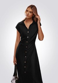 Vestido negro de botones con cuello, mangas cortas y una silueta ajustada. Presenta botones de madera en el frente y una textura suave.