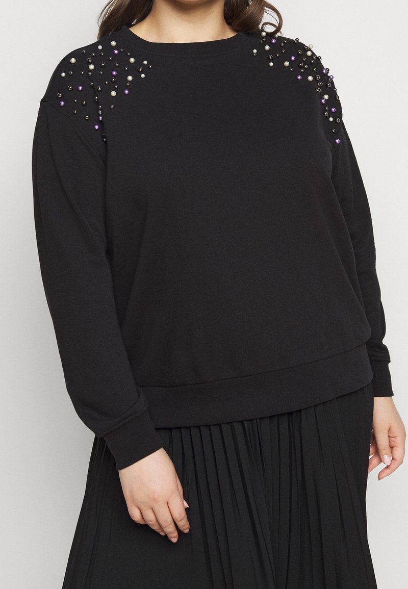 Haut noir à manches longues avec ornements en perles et en perles sur les épaules, assorti à une jupe noire plissée, porté par une personne aux cheveux lâchés.