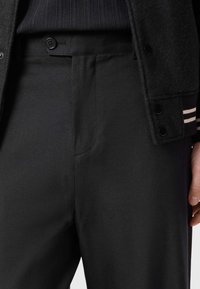 Zwarte broek van gladde stof met een getailleerde pasvorm. Voorzien van een voorzak en knoopsluiting, met een zichtbare, gestructureerde tailleband.