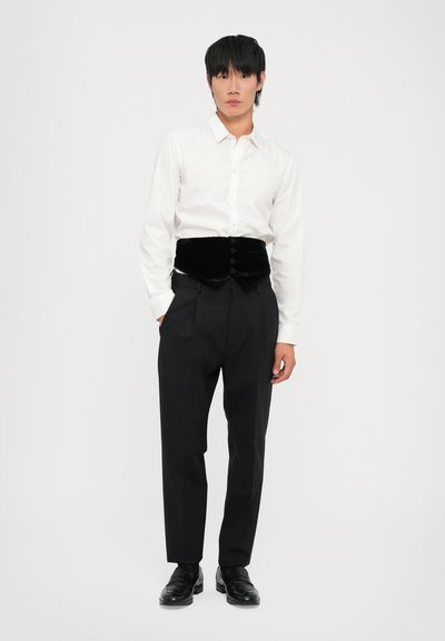 Weißes Hemd mit Knöpfen, schwarzes Samtwesten mit Knöpfen, schwarze Hose und schwarze Schuhe. Einfarbiger weißer Hintergrund.