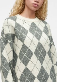 Jeune personne aux cheveux blonds bouclés portant un pull gris et blanc à motif losanges avec un col rond.
