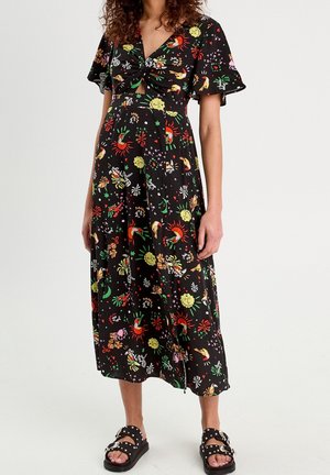 Femme portant une robe midi noire avec des motifs colorés de soleil, d'oiseau et d'étoile, et des sandales noires cloutées, debout sur un fond blanc.