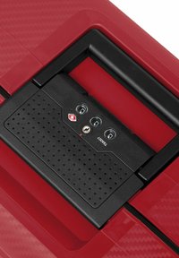 Valigia rossa con una finitura testurizzata. Presenta un lucchetto nero approvato dalla TSA con tre quadranti e una serratura a chiave. Maniglia liscia integrata nel design.