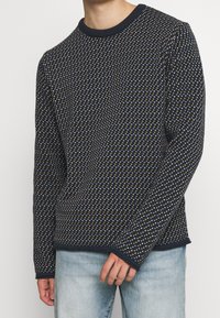 Stickad tröja i marinblått, med ett strukturerat mönster av små vita och gula former. Klassisk rund hals, långa ärmar, med ribbade muddar.