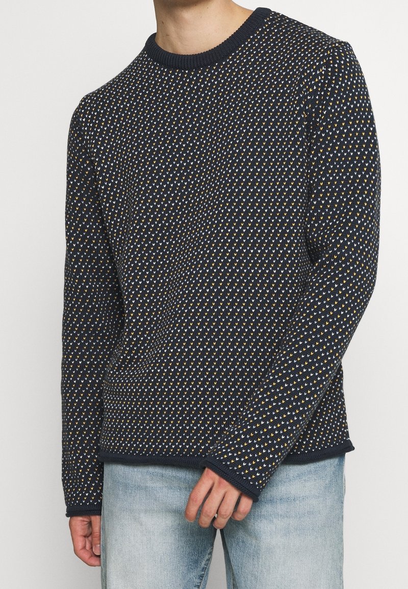 Stickad tröja i marinblått, med ett strukturerat mönster av små vita och gula former. Klassisk rund hals, långa ärmar, med ribbade muddar.