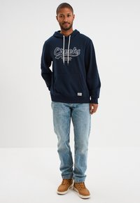 Sudadera azul marino con texto gráfico blanco, bolsillo frontal, cordón ajustable, combinada con jeans de un azul claro y zapatos beige.