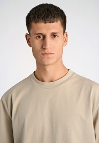 Vaaleanbeige, lyhythihainen t-paita, joka on valmistettu sileästä, kevyestä kankaasta, jossa on pyöreä pääntie ja hienovarainen rakenne. Mallilla on neutraali ilme.