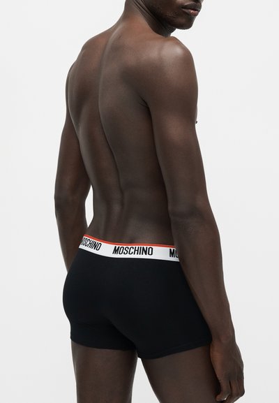 Άνδρας χωρίς πουκάμισο από πίσω, που φοράει μαύρα μπόξερ Moschino με λευκή μέση και κόκκινο περίγραμμα.