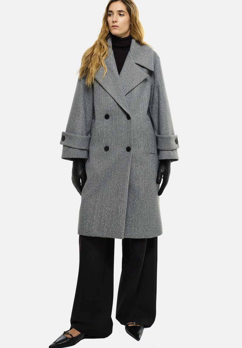 Manteau en laine gris avec fermeture croisée, grands revers et manches oversize. Porté sur un pantalon noir et des gants. Texture lisse.