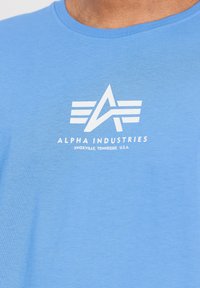 Svjetloplavi majica od mekog materijala s bijelim grafičkim logotipom kutnog dizajna i tekstom: "ALPHA INDUSTRIES, KNOXVILLE, TENNESSEE, S.A.D."