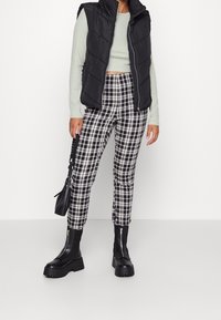 Personne portant un gilet matelassé noir sans manches, un crop top à manches longues vert clair, un pantalon à carreaux noir et blanc, des bottes noires épaisses, et tenant un sac à main noir.