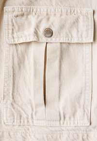 Beige denimkangas, jossa on rintatasku, jonka kannessa on läppä ja metallinen nappisuljenta. Kankaassa on teksturoitu pinta ja hienovarainen herringbone-kuvio.