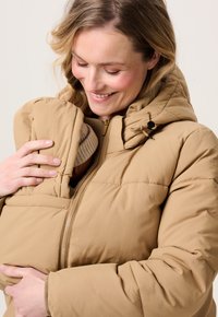 Een vrouw die naar beneden glimlacht terwijl ze een baby dichtbij houdt, beiden gekleed in bijpassende beige gewatteerde jassen, waarbij de baby gedeeltelijk door de jas is bedekt.