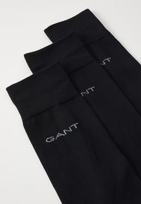 Svarte sokker med ribbet design og en logo brodert i lys grå. Tre par stablet, som viser oppbretten og materialets tekstur.