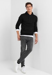 Zwarte hoodie met een voorzak, gecombineerd met grijze jeans en witte sneakers. Model staat tegen een lichtgrijze achtergrond.