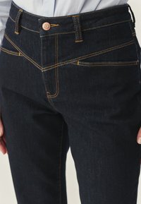 Jeans de mezclilla oscura con un diseño ajustado, que presentan costuras amarillas distintivas, un cierre de botón de cobre y bolsillos delanteros con corte en ángulo.