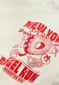 Biela tričko s červenou grafickou potlačou postavy šišky vedľa paradajky, s textom "NEW YORK BAGEL RUN" a "Čerstvé."