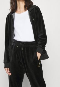 Žena v černé sametové zipové bundě a odpovídajících stahovacích kalhotách se zlatými akcenty, doplněná bílým ribovým topem bez rukávů.