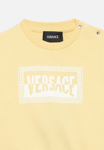 Gul Versace sweatshirt med hvid græsk nøglekantramme og logo tekst, med to sølv trykknapper på skulderen nær halsudskæringen.