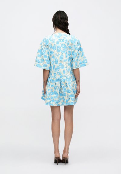 Γυναίκα στέκεται με την πλάτη, φορώντας κοντό μπλε και λευκό φόρεμα με λουλούδια, φαρδιά μανίκια και βολάν στο τελείωμα, σε συνδυασμό με καφέ ψηλοτάκουνα παπούτσια.