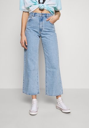 Femme portant un jean large bleu clair, des baskets blanches montantes et un haut court tie-dye, une main dans la poche.