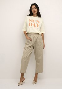 T-shirt en coton blanc avec le texte orange "SUN DAY Chill out", associé à un pantalon large à taille haute beige et des chaussures slip-on beiges.