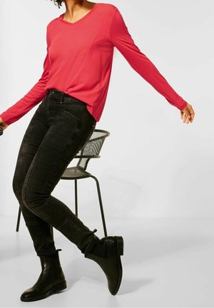 Rotes langärmliges V-Ausschnitt-Shirt kombiniert mit schwarzen Skinny Jeans und Stiefeletten. Schwarzer Stuhl im Hintergrund. Sauberer, minimalistischer Stil.