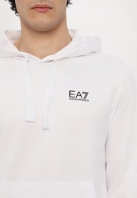 Hvid hoodie lavet af blødt stof, har en lomme foran, dråbesnor til hætten og et sort EA7 Emporio Armani-logo på venstre bryst.