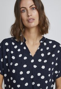 Granatowa bluzka w białe kropki. Posiada dekolt w kształcie litery V, krótkie rękawy oraz fakturowany materiał z subtelnymi wypukłymi wzorami.