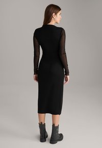 Schwarzes Midikleid mit langen transparenten Ärmeln, figurbetontem Design und glatter Textur. Kombiniert mit schwarzen Knöchelstiefeln mit einer klobigen Sohle.