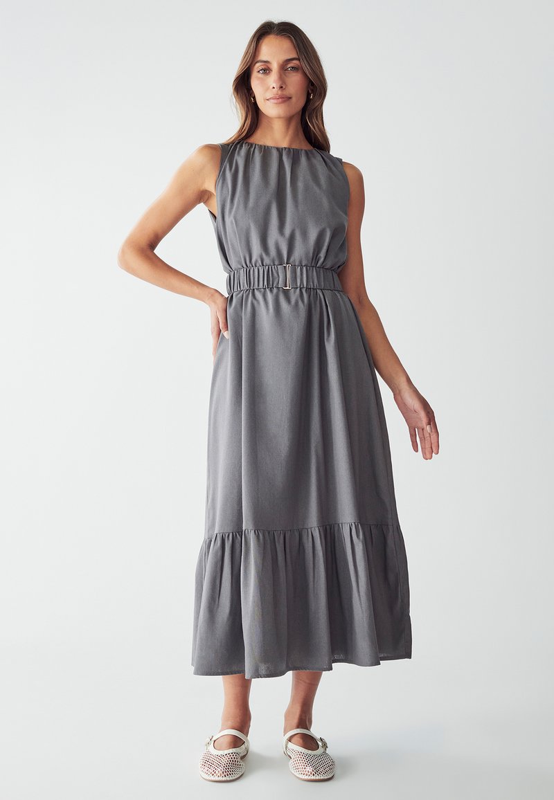 Vestido maxi sin mangas de color gris con escote fruncido, cintura elástica y dobladillo con volantes en capas. Llevado con zapatos de slip-on de color claro.