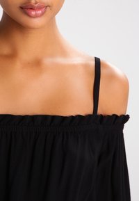 Top nero off-shoulder con bordi arricciati, caratterizzato da spalline sottili e una consistenza morbida, che mostra una vestibilità rilassata attorno alle spalle.