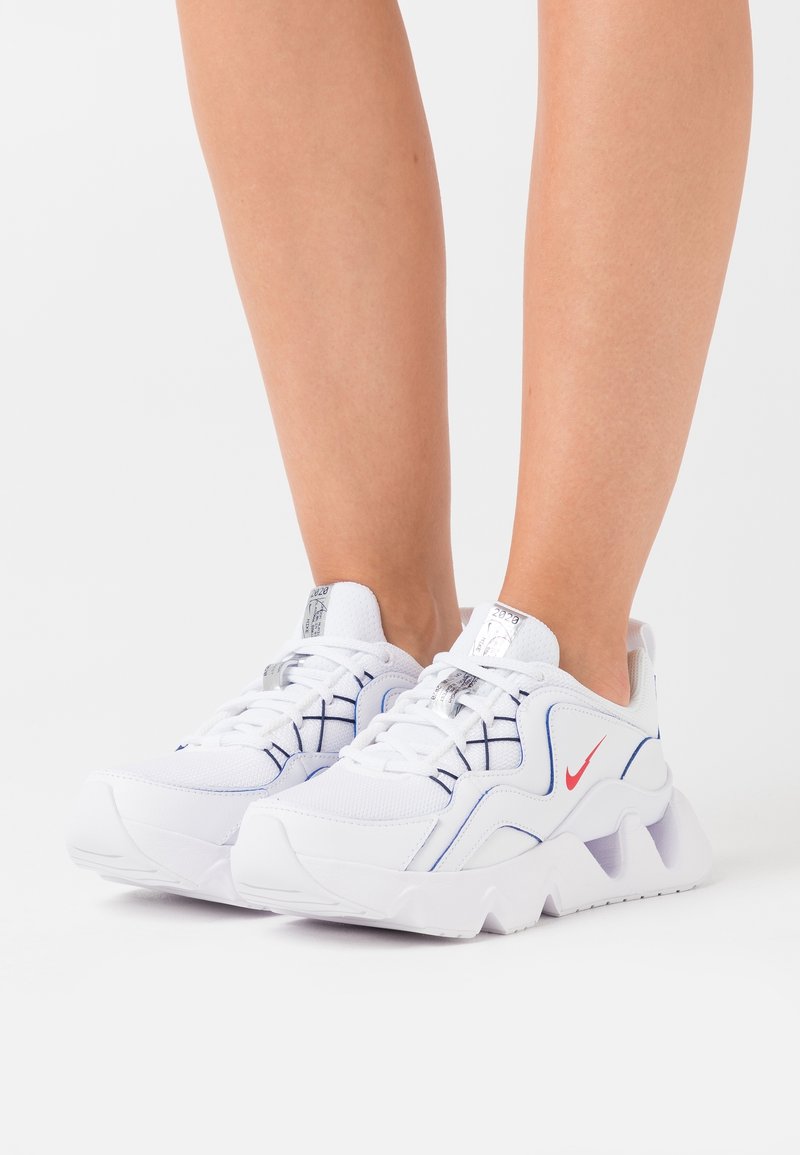 Baskets blanches de sport avec tige en mesh texturé, accents bleus et logo rouge. Semelle extérieure blanche épaisse avec détails ajourés. Design à lacets.