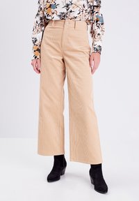 Pantalon large en velours côtelé beige à taille haute, avec une fermeture à un bouton et une texture côtelée discrète, porté avec des bottines noires.