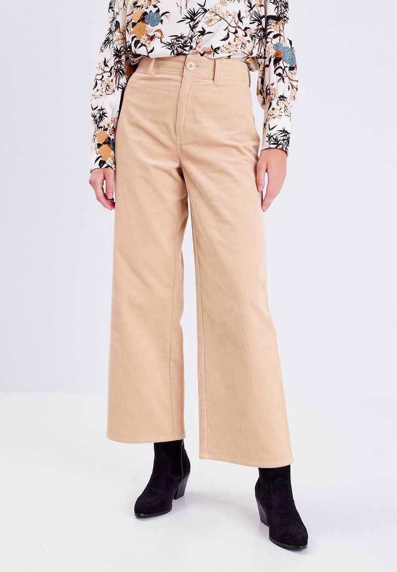 Pantalon large en velours côtelé beige à taille haute, avec une fermeture à un bouton et une texture côtelée discrète, porté avec des bottines noires.