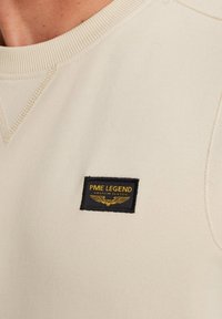 Beige Sweatshirt mit einem gerippten Rundhalsausschnitt und einem schwarzen rechteckigen Patch mit goldener Schrift und Flügel-Logo-Detail.