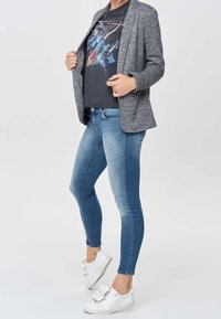 Blazer texturizado gris sobre una camiseta gráfica negra, combinado con jeans ajustados azul y zapatillas blancas con un detalle de lazo en los cordones.