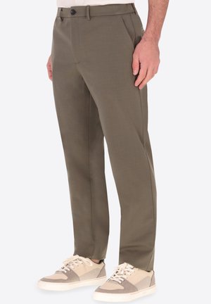 Uomo che indossa pantaloni color verde oliva a gamba dritta e sneakers beige in piedi con la mano destra rilassata lungo il fianco.