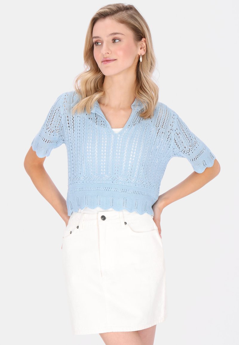 Młoda kobieta o falowanych blond włosach, ubrana w jasnoniebieski dzianinowy top z krótkim rękawem i białą spódnicę z wysokim stanem, z rękami na biodrach.