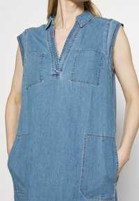 Abito in denim con colletto, maniche corte, due tasche frontali e vestibilità comoda. Il tessuto è di un azzurro chiaro, con una texture lavata.