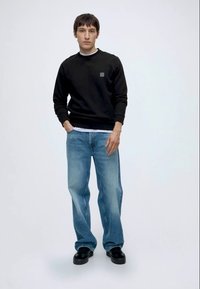Jeune homme debout portant un sweat-shirt noir à manches longues, un jean ample bleu clair et des chaussures noires sur un fond blanc uni.