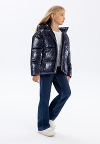 Giubbotto imbottito blu navy con finitura lucida, cappuccio, bottoni a scatto dorati e design trapuntato. Abbinato a jeans scuri e sneaker bianche.