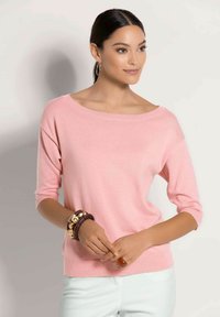 Frau mit dunklen Haaren, die einen pinkfarbenen Pullover mit Dreiviertelärmeln, weiße Hosen, Creolen, holz- und goldfarbene Armbänder sowie einen roten Ring trägt.