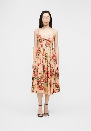 Frau steht und trägt ein ärmelloses, pfirsichfarbenes Midikleid mit rotem und pinkfarbenem Blumenmuster sowie schwarze High-Heel-Sandalen mit Knöchelriemen.