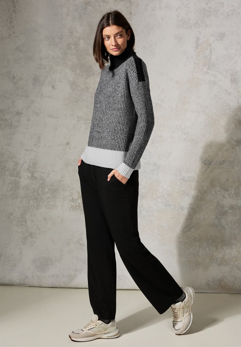 Pull en tricot gris et noir avec un col montant et un ourlet rayé, associé à un pantalon noir et des baskets légères. Texture lisse et coupe décontractée.