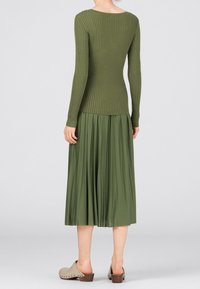 Oliivinvihreä ribbipintainen pitkähihainen paita, jossa on pyöreä kaula-aukko, yhdistetty pleated-midi-hameeseen, joka on samansävyinen, päällä beige klompi vaalealla taustalla.