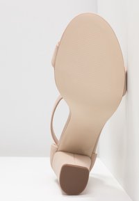 Sandale à talon haut nude avec un talon bloc, bout ouvert et fine sangle de cheville. Tige en synthétique lisse et semelle texturée pour une bonne adhérence.