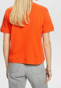 Oranssi puuvillainen t-paita, jossa on lyhyet hihat, rento istuvuus ja resorit hihansuissa. Yhdistetty vaaleanharmaisiin farkkuihin. Sileä pinta, ei kuvioita.