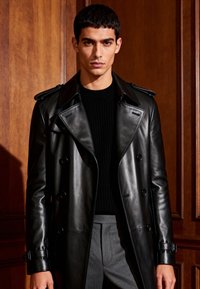 Double-breasted black leather trench coat with shoulder epaulets and buckle details. Worn over a black ribbed sweater, paired with gray trousers.