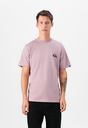 Hellblaues Baumwoll-T-Shirt mit rundem Halsausschnitt, kurzen Ärmeln und einem schwarzen Logo-Patch auf der linken Brust. Es werden schwarze Hosen getragen.